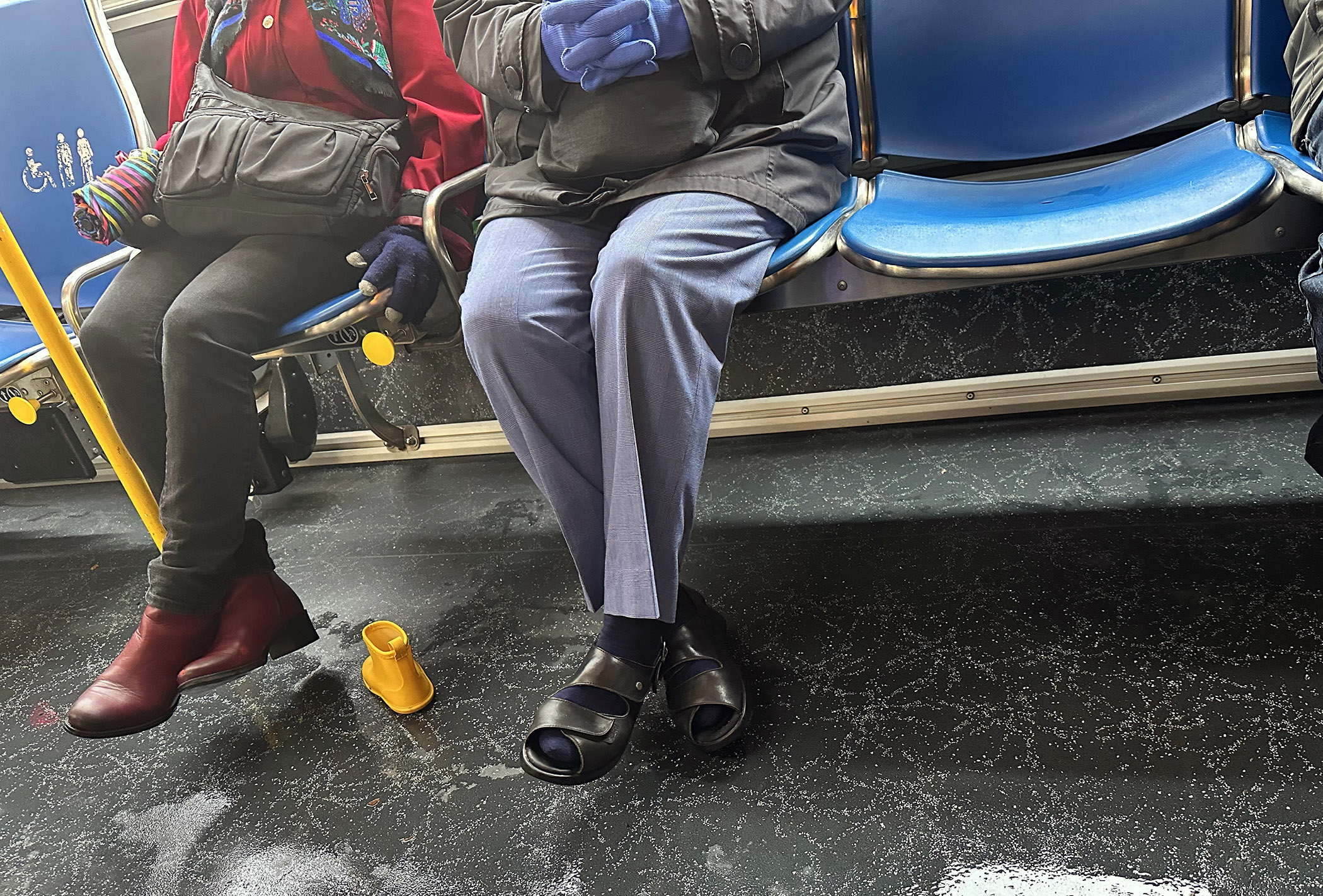 Two people sit on a bus; one is wearing red boots, and the other is in black sandals with blue pants. A yellow rubber boot is on the floor.
