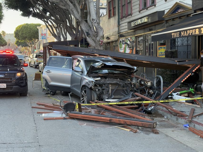 Car crashed into an outdoor dining structure on a city street, with debris scattered and police vehicle nearby.