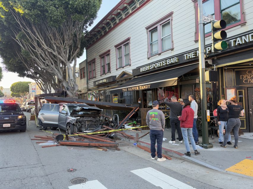 Car crash into a building; silver car damaged under a nearby tree and debris scattered. Several people observe the scene while police vehicle is parked in the street.
