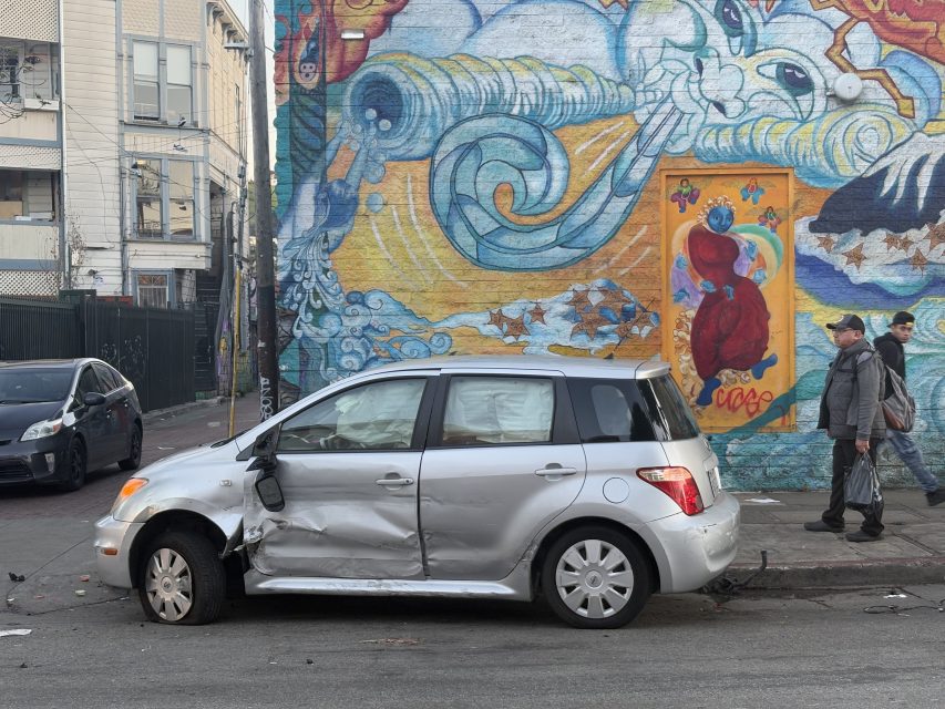 A silver car with severe dents on its side, possibly from a recent police chase, is parked on a street. A colorful mural with abstract designs and figures covers the wall behind it as a person walks casually on the sidewalk.