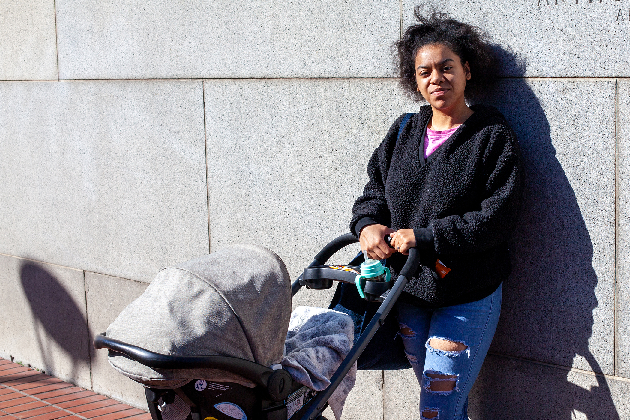 Person in a black jacket leans against a concrete wall, next to a baby stroller on a sunny day.