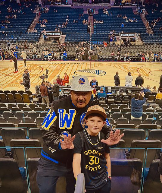 Two people posing for a photo in front of a basketball court. The man is wearing a Warriors jacket and cap, while the child is wearing a Golden State shirt and hat. Spectators are in the background.