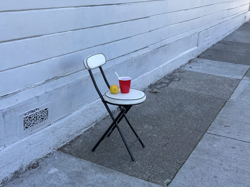 A white folding chair with a red cup, a lemon, and a spoon is placed on it against a white building wall on a sidewalk corner.