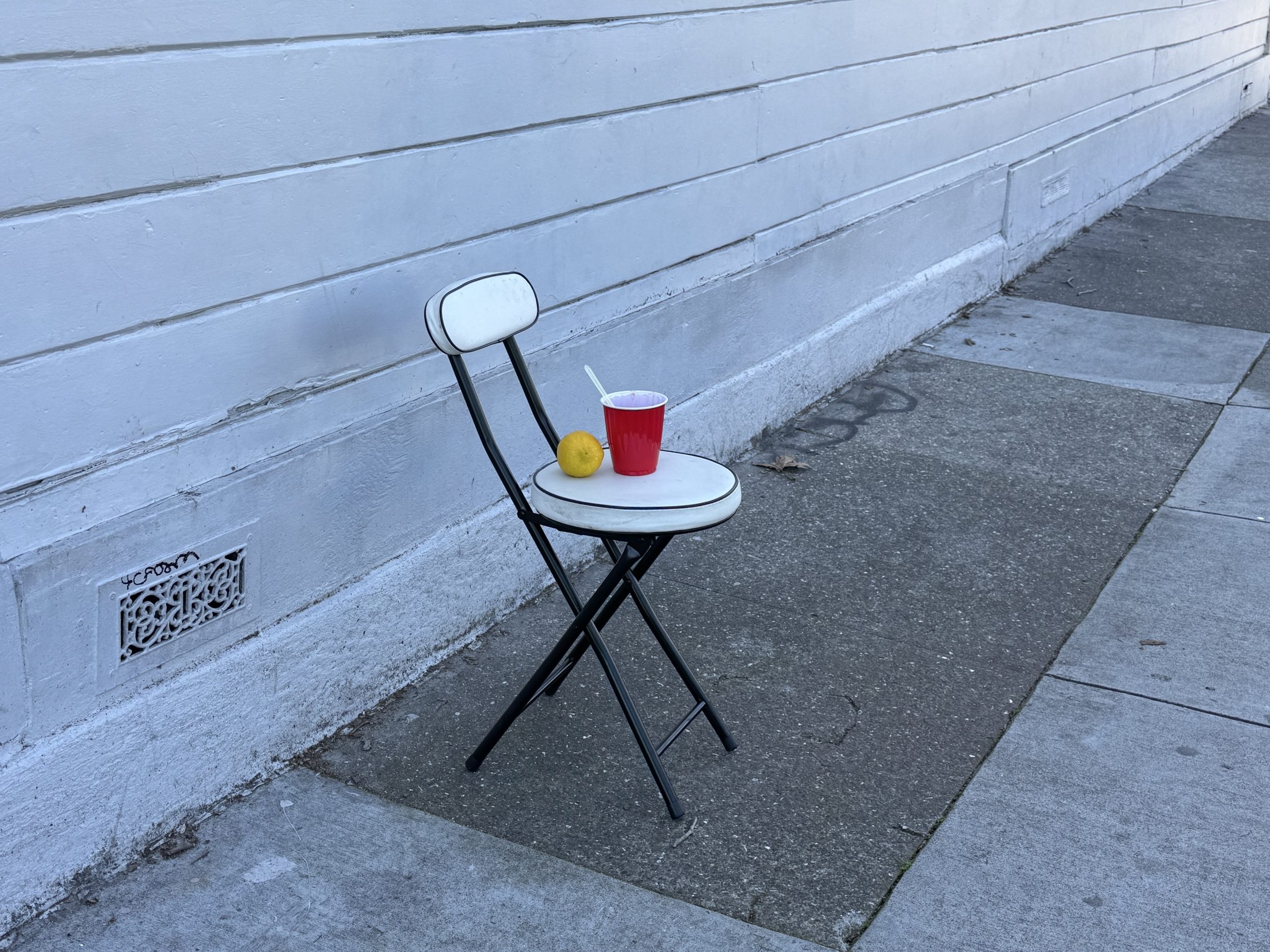 A folded white chair with a red cup, a spoon, and a lemon on the seat is placed against a white wall on a concrete sidewalk.