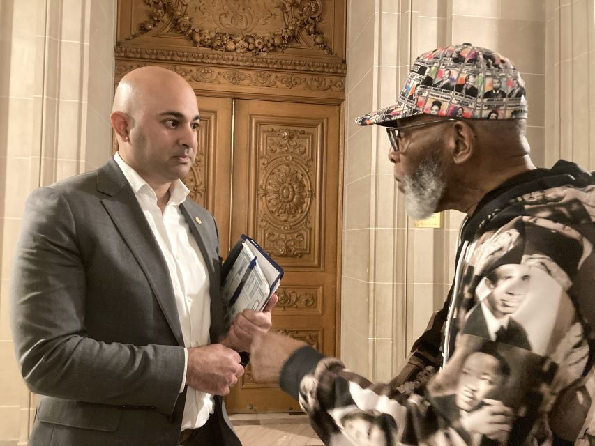 Two men are engaged in conversation in front of an ornate wooden door. One is in a suit holding papers, and the other is wearing a cap and patterned sweatshirt.
