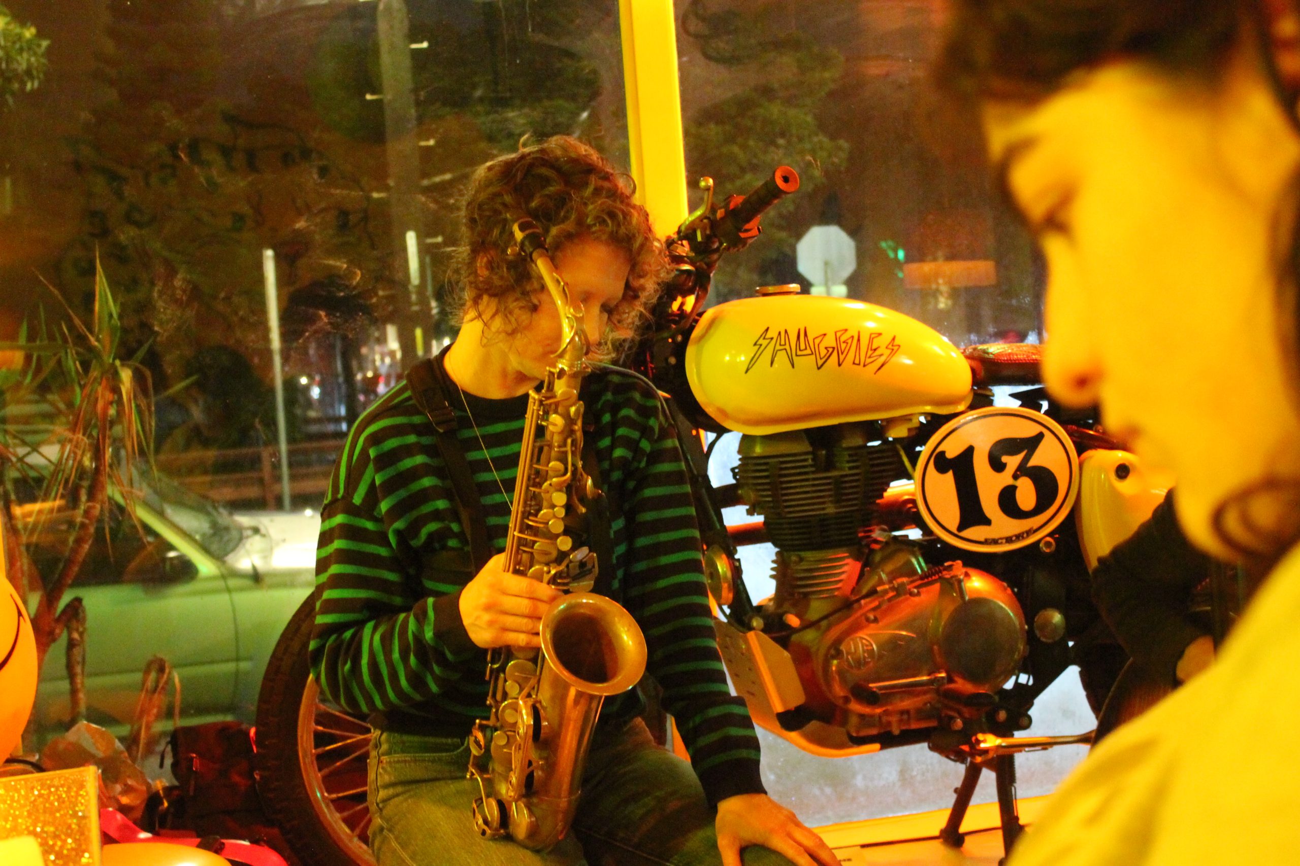 Person playing a saxophone next to a vintage motorcycle with "13" and "Saugles" written on it, inside a dimly lit room.