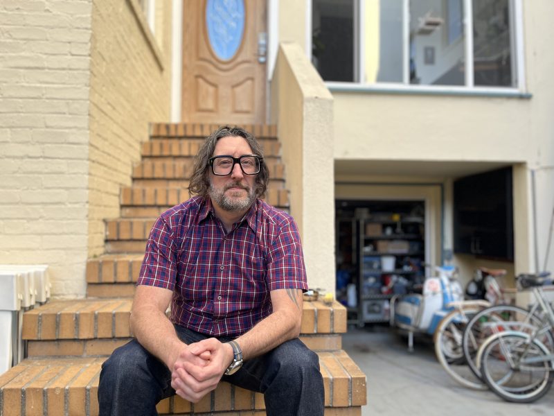 A man with glasses and long hair sits on brick steps, wearing a red plaid shirt. A house with an open garage full of items is in the background.