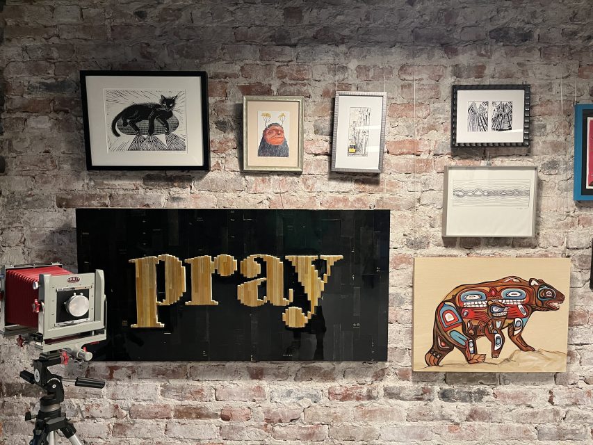 An art display on a brick wall includes a red vintage camera, a large "pray" sign, and various framed artworks, including animal-themed prints.