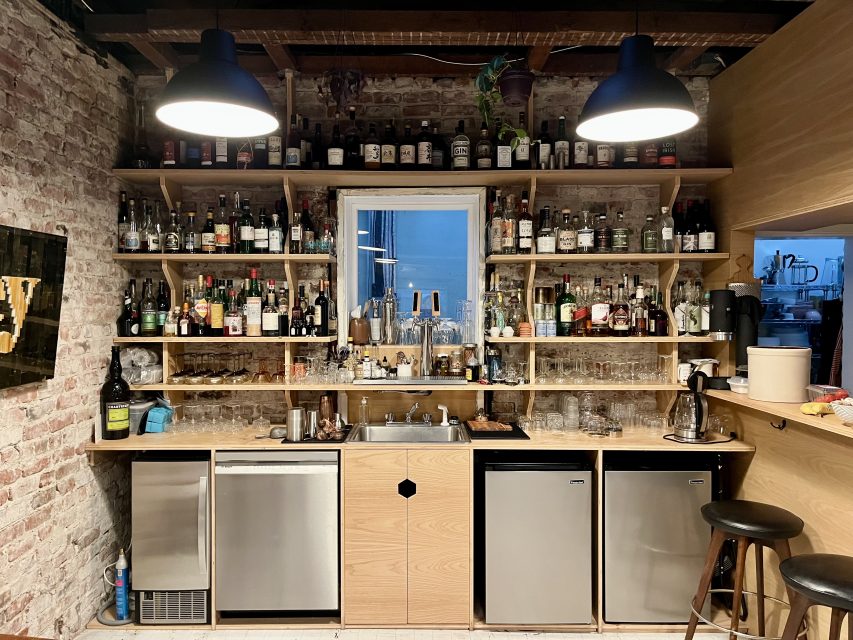 A home bar with shelves holding various bottles of liquor and glasses, a sink and mini fridges below, and two overhead lights illuminating the area.