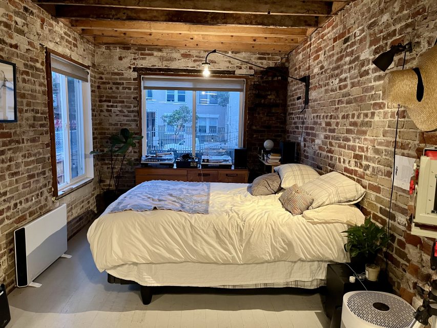 A cozy bedroom with exposed brick walls, a large window, a bed with pillows and a comforter, a small plant, and some electronics on a wooden chest of drawers.