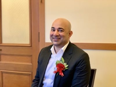 Bilal Mahmood in a suit with a red and white corsage sits in front of a wooden door, smiling at the camera.