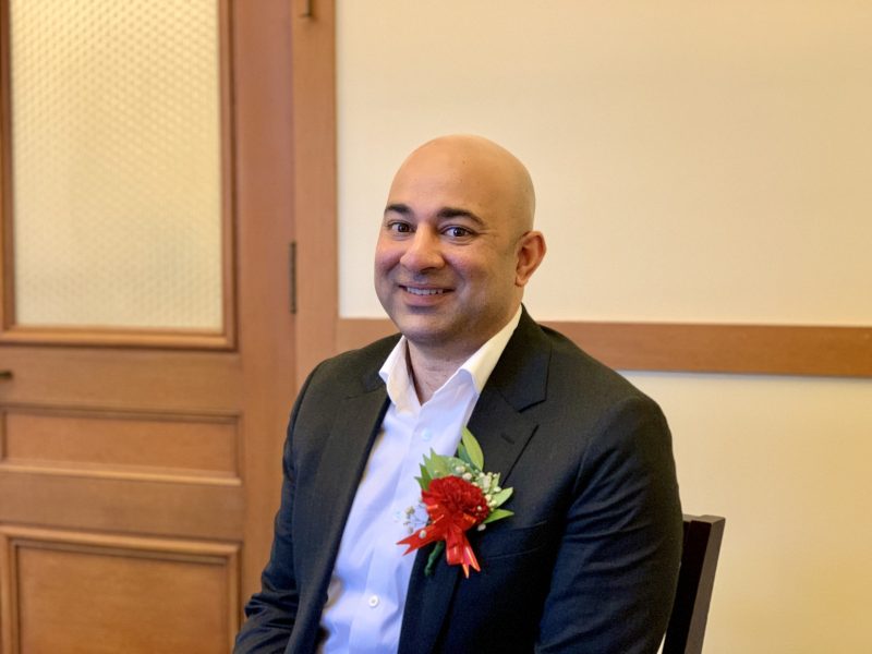Bilal Mahmood in a suit with a red and white corsage sits in front of a wooden door, smiling at the camera.