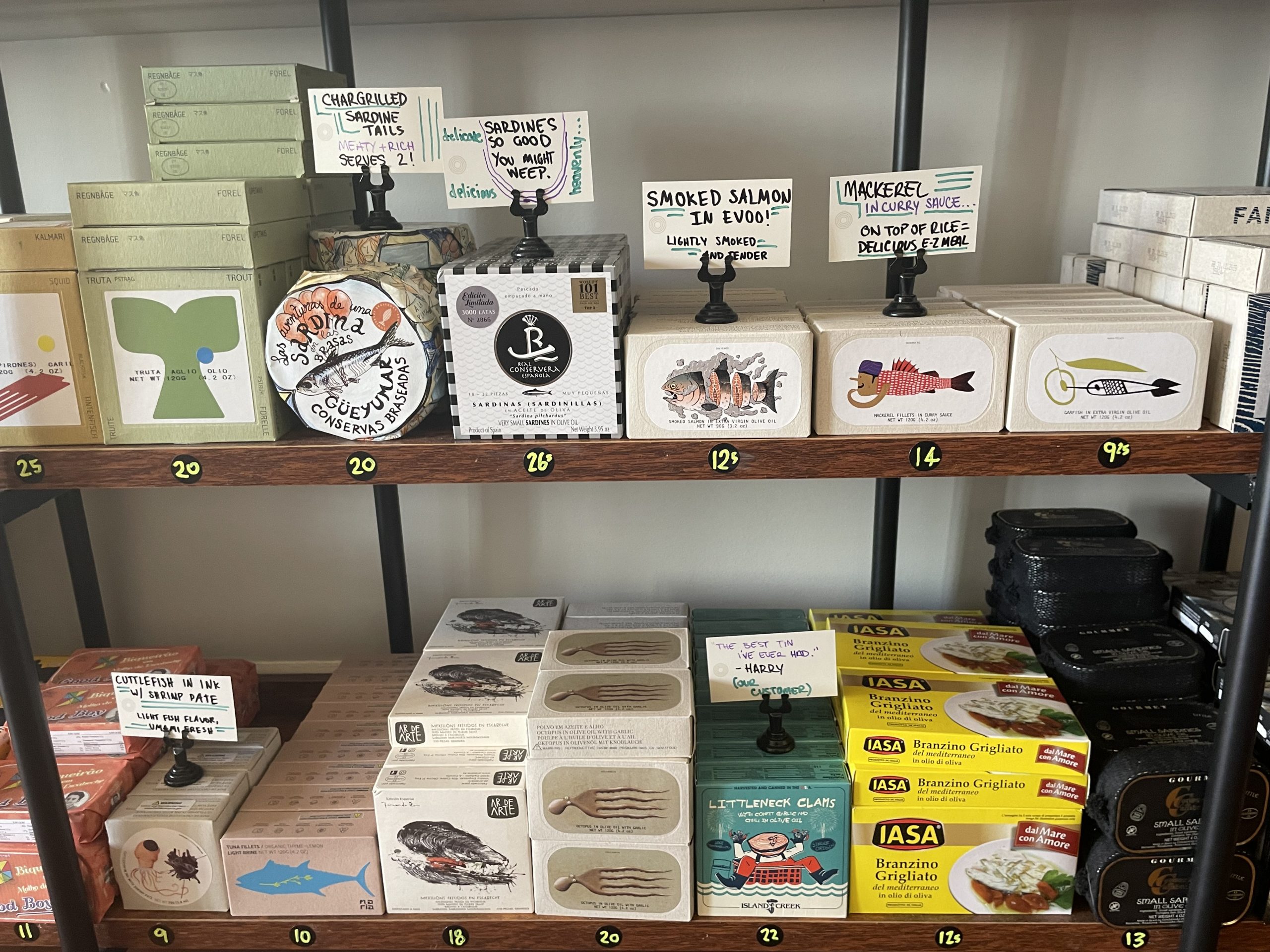 Shelves displaying a variety of seafood cans and boxes, including sardines, smoked salmon, and mackerel, with handwritten signs indicating prices and product details.