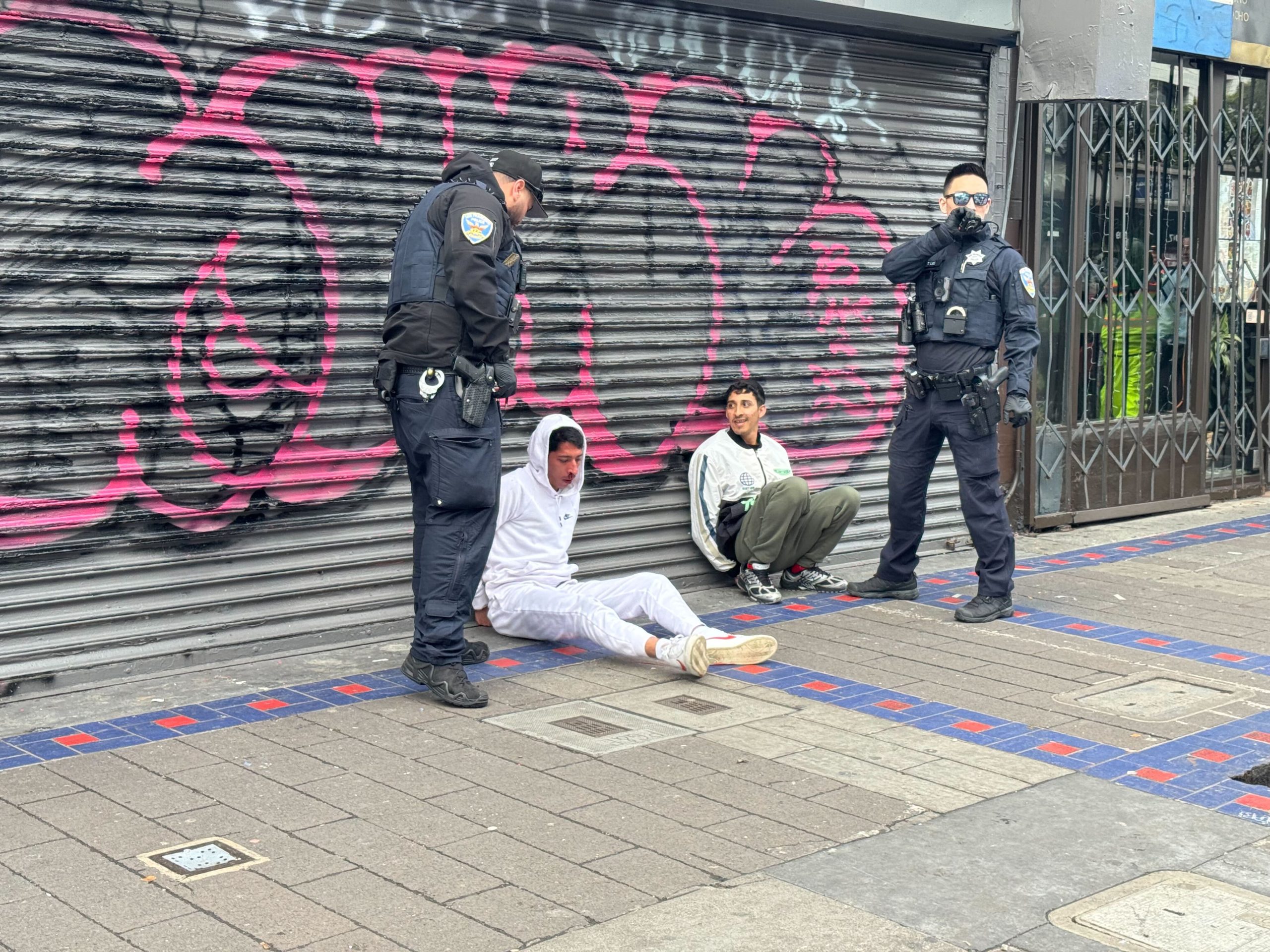 Two police officers stand near two seated individuals in front of a graffiti-covered shutter on a city sidewalk.