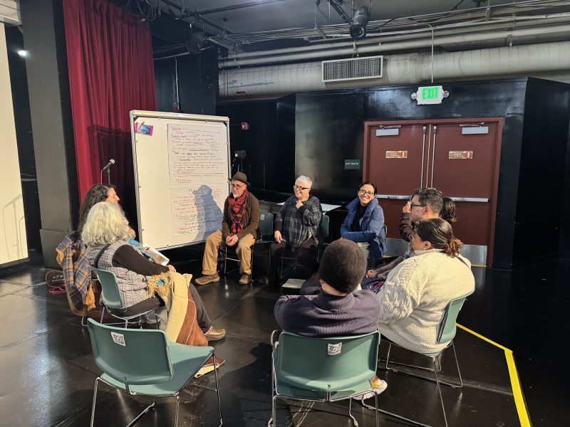 A diverse group of eight people sit in a circle on chairs in a room, engaged in discussion, with a whiteboard and flip chart in the background.
