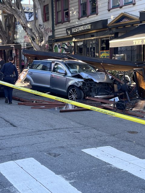 A silver car crashes into an outdoor dining area, surrounded by debris and police tape, with officers and a restaurant in the background.