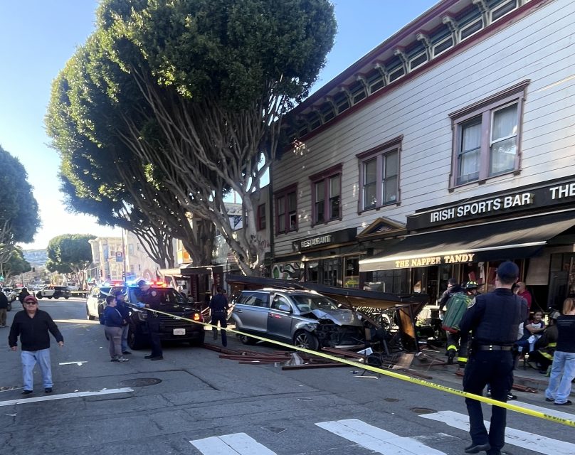 A car involved in a police chase has crashed onto the sidewalk in front of a building with "Irish Sports Bar" signage. Police and onlookers are at the scene, surrounded by yellow caution tape.