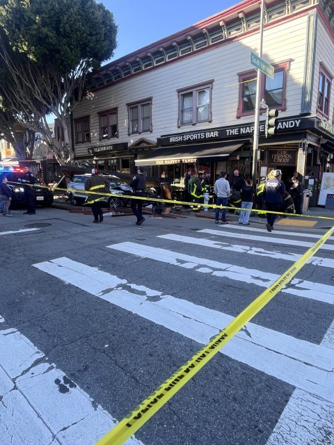 A police scene outside an Irish sports bar with yellow caution tape. Officers and bystanders stand near a parked black car on a street corner.