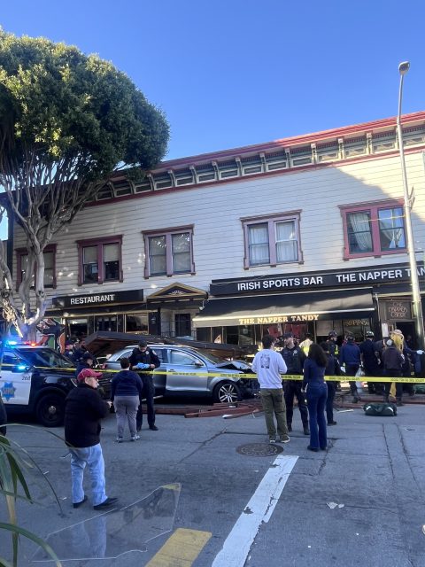 A car has crashed into the front of a building labeled "Irish Sports Bar." Several bystanders and police officers are present, with police tape cordoning off the area.