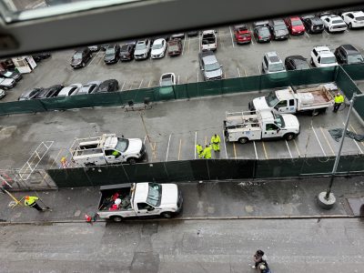 Aerial view of a parking lot with several white utility trucks and workers in high-visibility clothing. A green temporary fence separates the lot from parked cars.