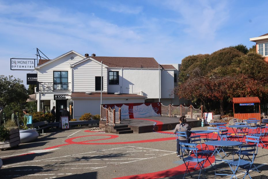 Outdoor seating area with blue chairs and tables on a patterned pavement. A white building with signs for an optometry office and "XCOR" is in the background. Trees are on the right.