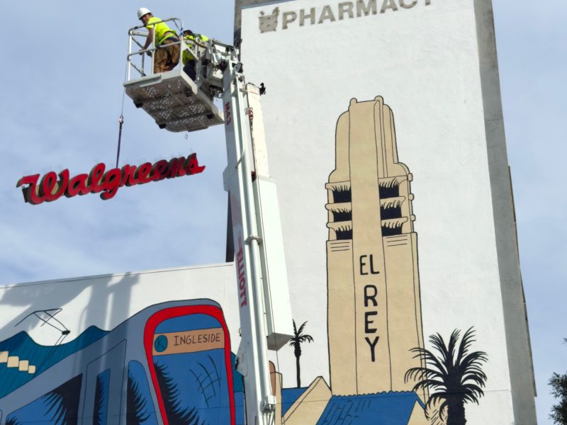 Workers on a lift remove a Walgreens sign on a building with a large mural of El Rey Theatre and "PHARMACY" text at the top.