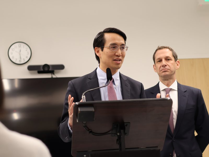A person speaking at a podium with a microphone, wearing a suit and tie. Another person stands behind, also in a suit. A wall clock and TV are in the background.
