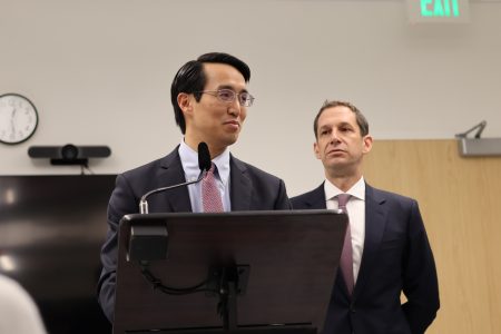 Two men in suits stand at a podium in an office setting. One speaks into a microphone while the other listens. A clock, television, and exit sign are visible in the background.