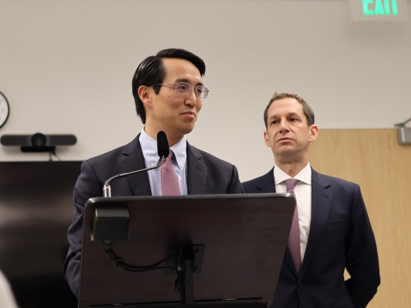 Two men in suits stand at a podium in an office setting. One speaks into a microphone while the other listens. A clock, television, and exit sign are visible in the background.