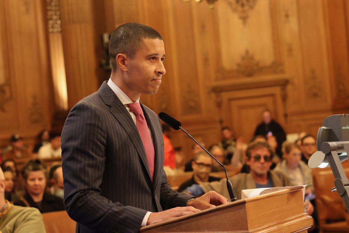 A person in a suit and tie speaks at a podium in a formal, wood-paneled room. Audience members sit in the background.