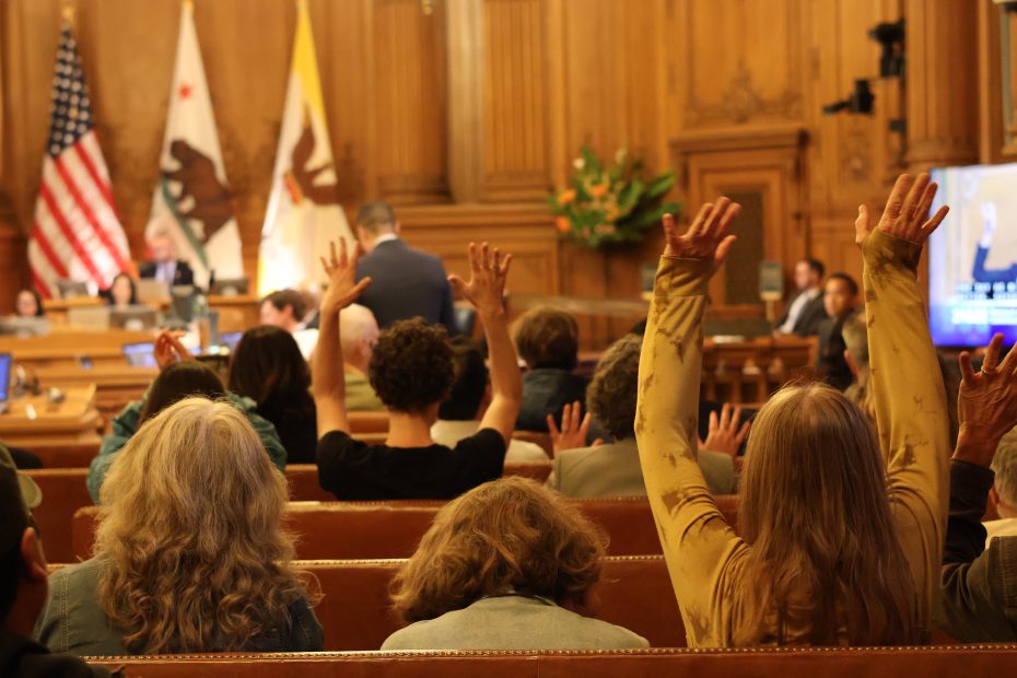 Audience members raise their hands during a formal meeting in a wood-paneled room with flags and a speaker at the front.