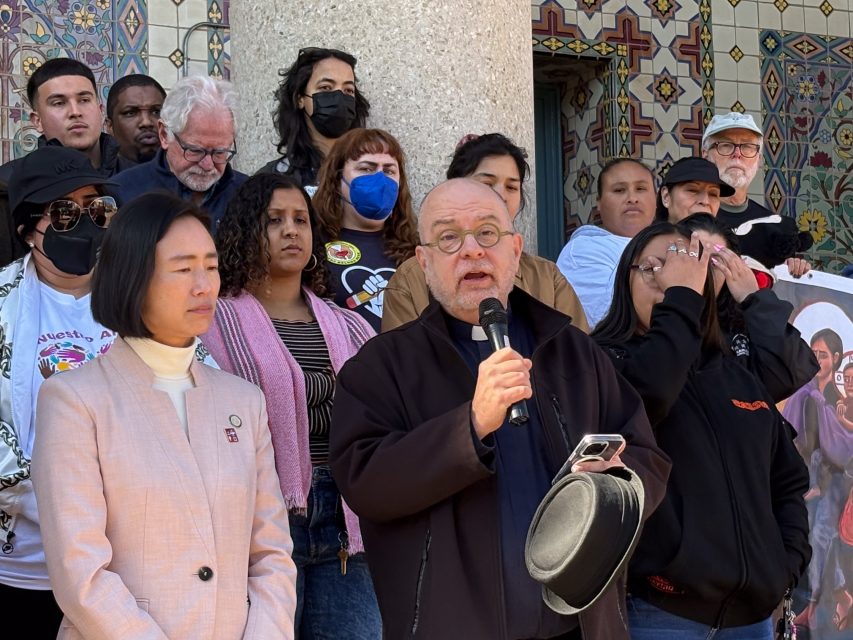 A group of people stands together in front of a decorated building; a man speaks into a microphone while others look on.