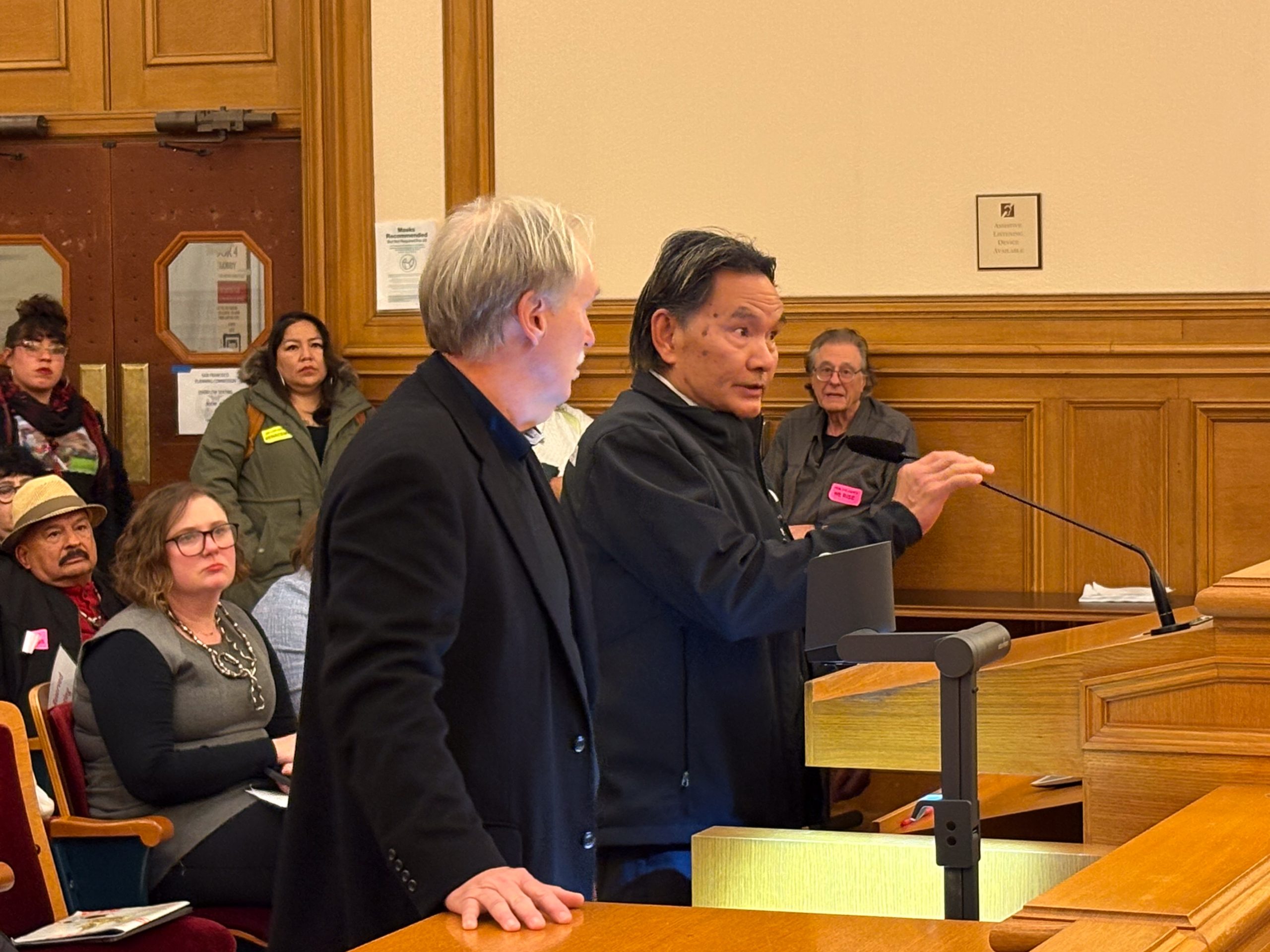 Two men stand at a podium speaking to an audience in a wood-paneled room. Several people are seated in the background, watching them.