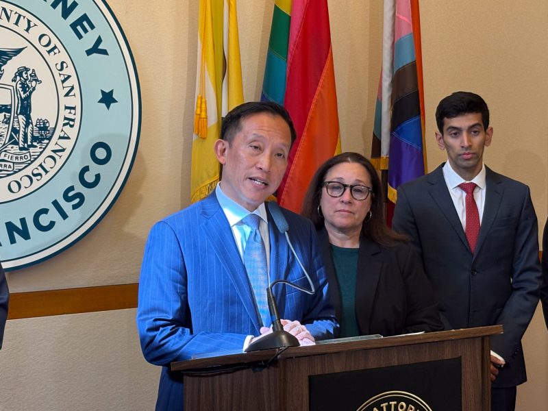A person in a blue suit speaks at a podium with two others standing nearby. A San Francisco seal and various flags are in the background.