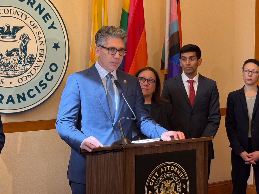 A man in a blue suit speaks at a podium in front of a City Attorney seal, accompanied by three people in formal attire. Flags are displayed in the background.