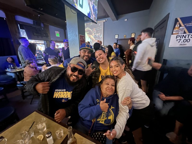 A group of people at a bar poses together, smiling. Many are wearing basketball-themed clothing. Drinks and a menu are on the table in front of them.