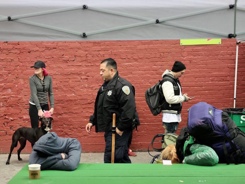 People at an outdoor area: a woman with a dog, an officer walking, a person texting, and another resting on a green table under a canopy. A brick wall serves as the background.