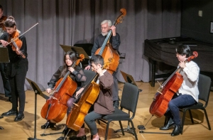 A string ensemble performing on stage, including a violinist, two cellists, and a double bassist, with music stands in front of them. A piano is visible in the background.