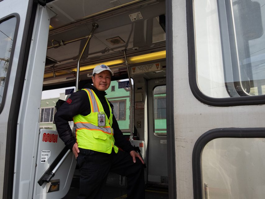 Person wearing a safety vest and cap stands at the entrance of a transit vehicle, smiling. 