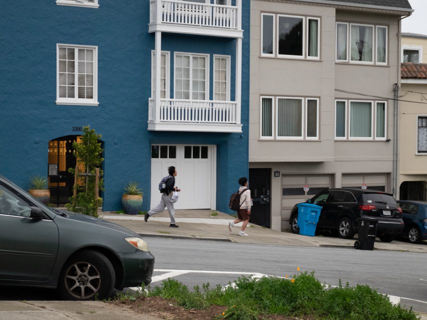 Two people walk on a sidewalk next to a blue building with balconies. A car is parked on the street, and trash bins are visible in front of a gray building.