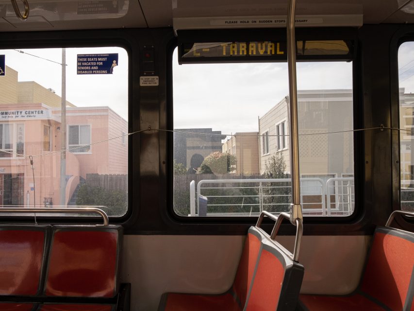 Interior of a bus with red seats, showing a view through the window of buildings on a street. Signs above the window indicate the destination and safety instructions.