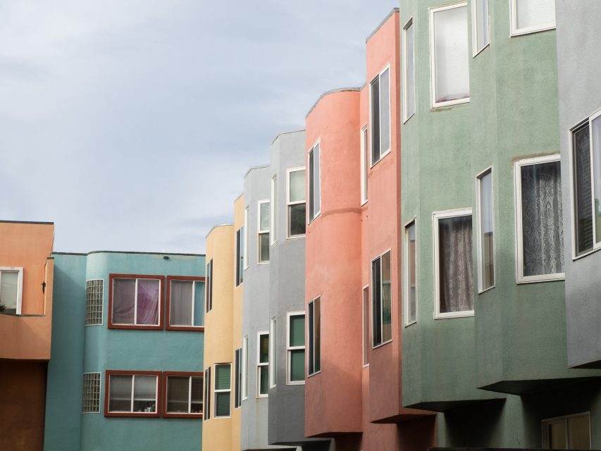 Row of colorful, geometrically-styled apartment buildings featuring pastel shades of blue, orange, green, pink, and gray under a cloudy sky.