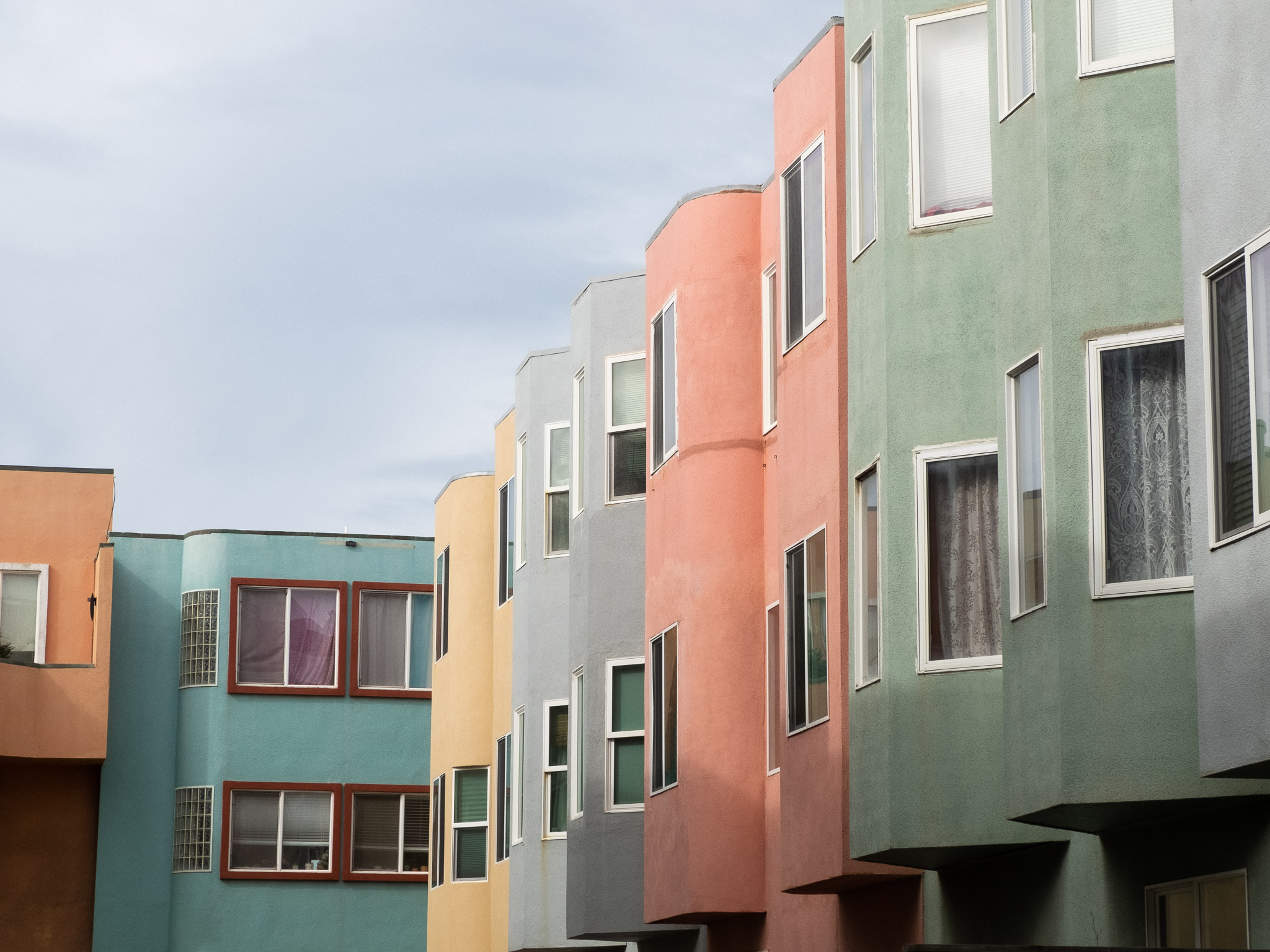 Row of colorful, geometrically-styled apartment buildings featuring pastel shades of blue, orange, green, pink, and gray under a cloudy sky.