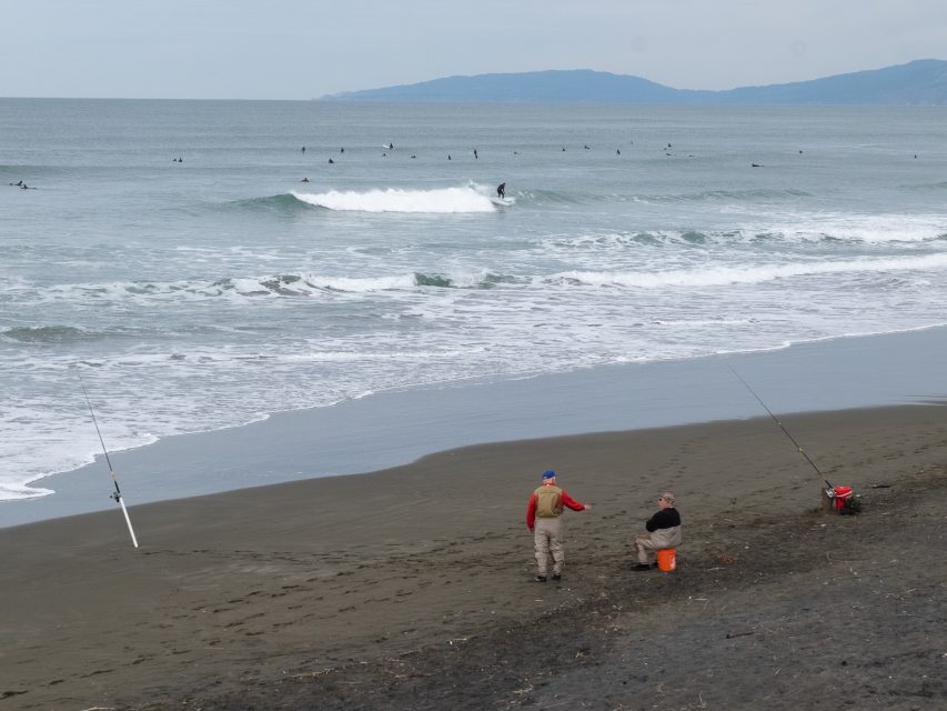 Two people fishing on a sandy beach with a fishing rod planted in the sand. Surfers are visible in the ocean waves. Distant land is visible across the water.