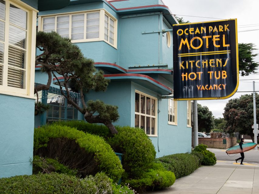 A teal building houses the Ocean Park Motel. The sign advertises kitchens, hot tubs, and vacancy. A person with a surfboard walks on the sidewalk.