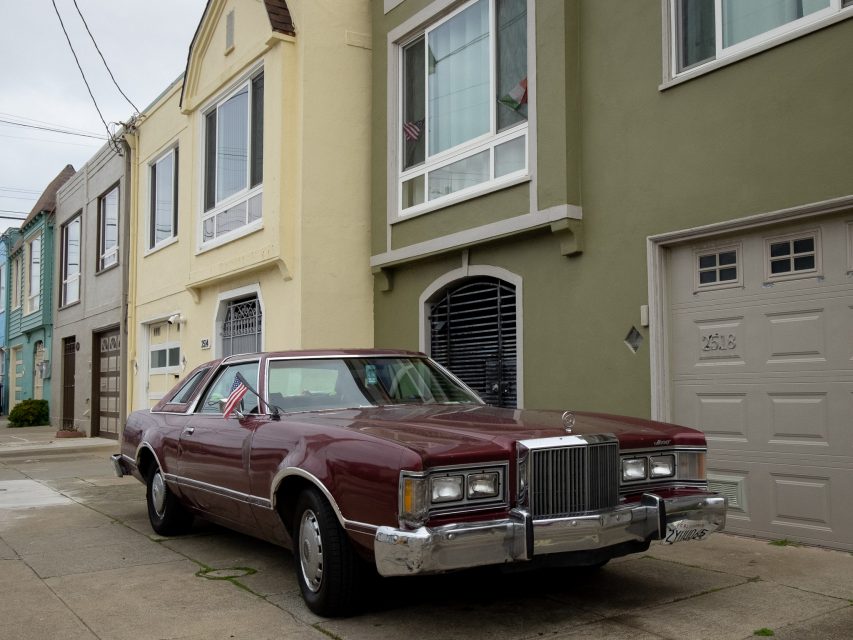 A vintage maroon car parked on a residential street in front of pastel-colored houses, with small flags on the windows.