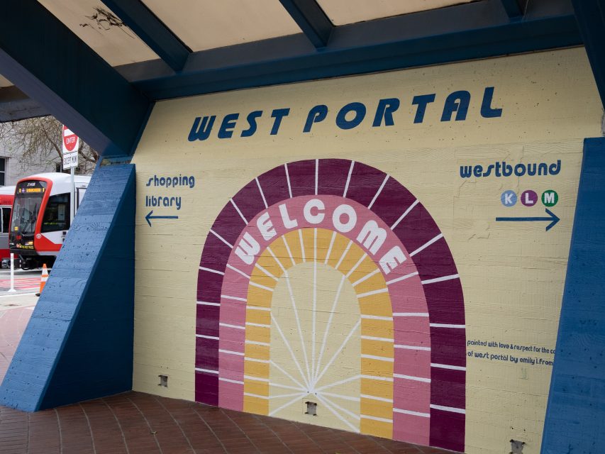 Colorful mural at West Portal station entrance with "Welcome" inside an arch, and directional arrows to shopping, library, K, L, M lines. Train visible on street in the background.