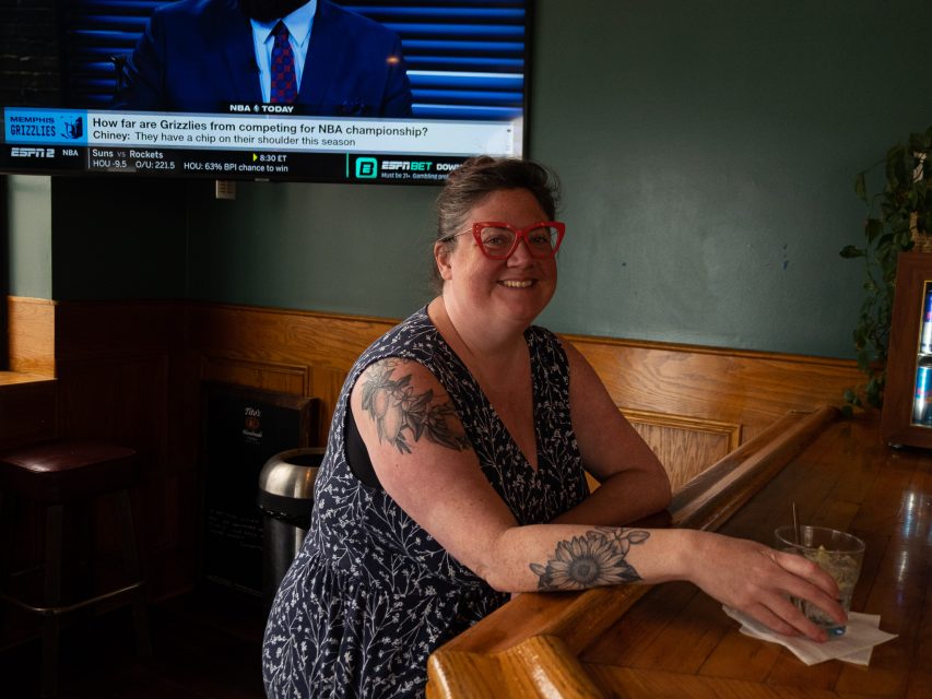 A person with red glasses and tattoos smiles while holding a drink at a bar. A TV with sports content is on in the background.