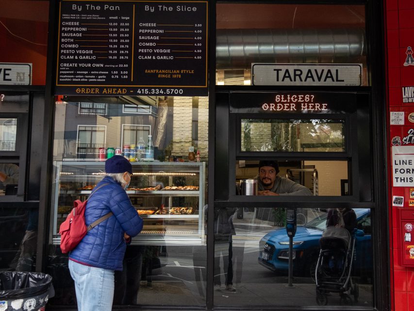 A woman in a blue jacket stands at a pizza shop window labeled "Taraval," looking at a menu board, while a man inside serves her coffee.