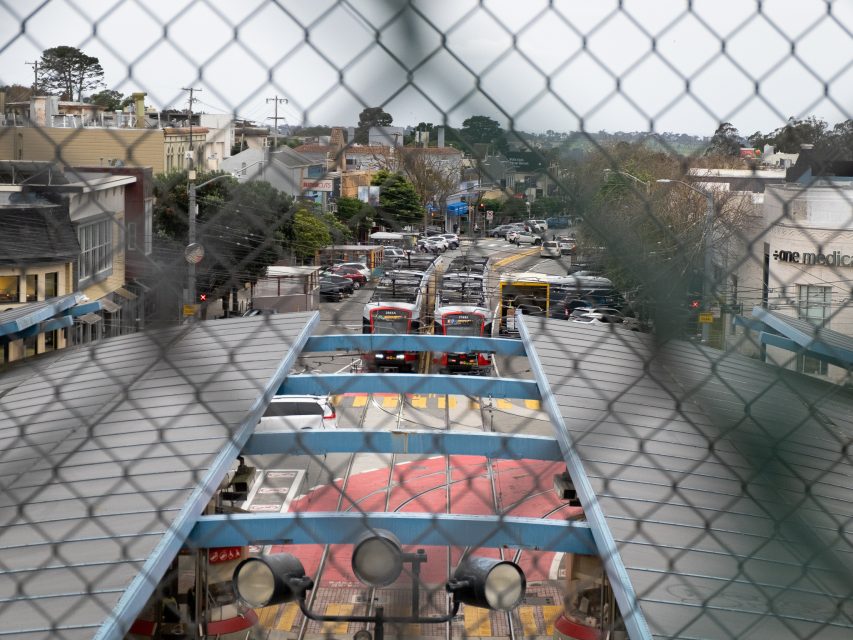 View through a chain-link fence of a busy urban intersection with cars, buses, and buildings lining the street.